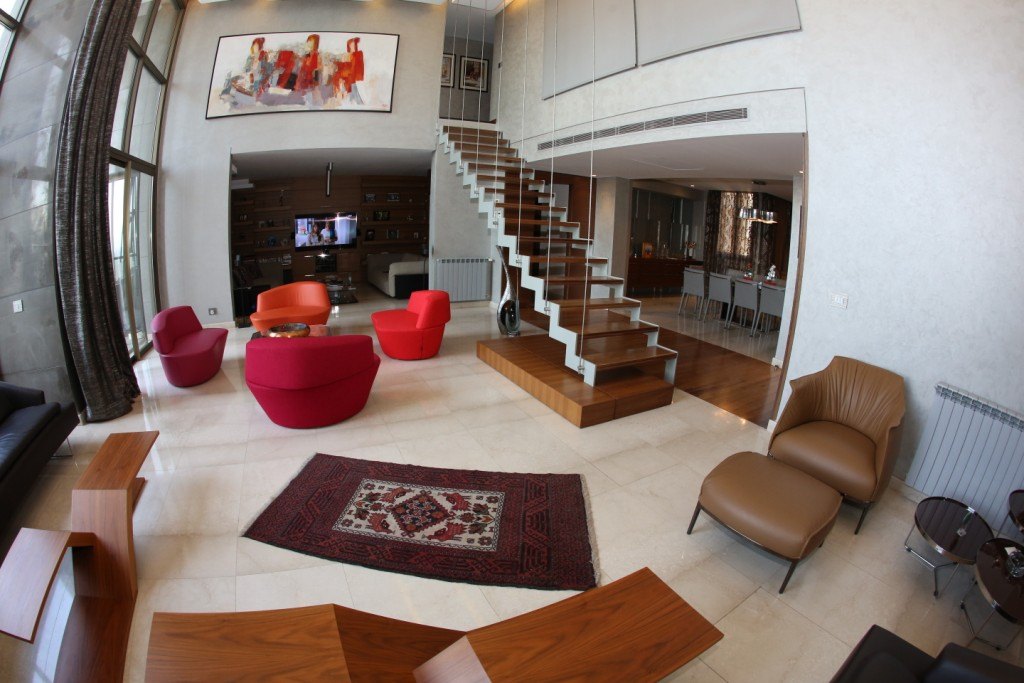 Séjour en double hauteur avec fauteuils rouges, tapis persan et escalier en noyer suspendu, marbre clair au sol.