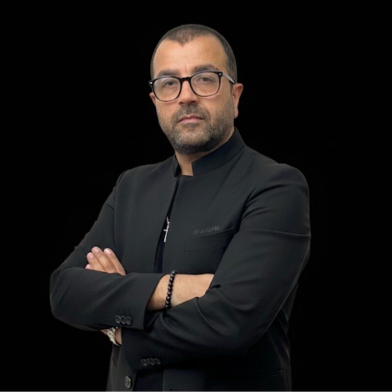 Portrait de Joseph I. Mouawad, bras croisés, veste sombre sur fond noir.